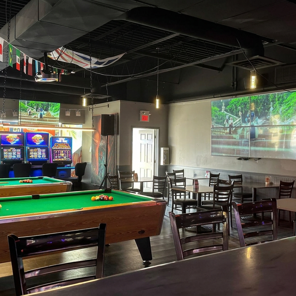 Sports bar interior with two pool tables, gaming machines, wall-mounted screens, international flags, and restaurant seating.